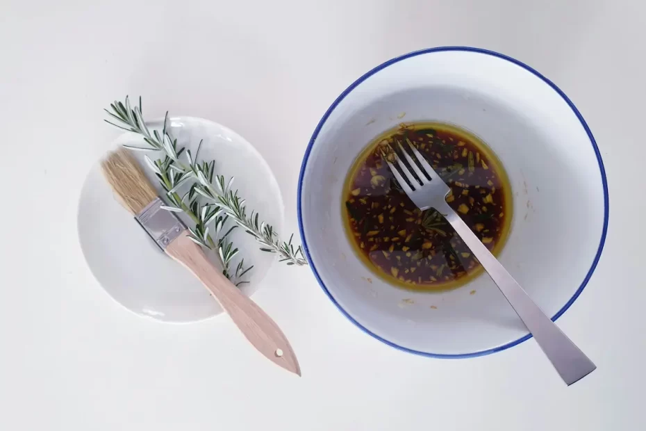 A bowl of dark brown marinade with herbs and garlic cloves, with a fork resting in it. Next to the bowl is a small white plate with a basting brush and a sprig of rosemary. All items are on a white surface.