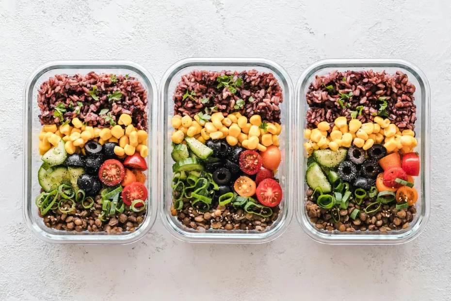 Three rectangular glass meal prep containers, each filled with layers of food. From top to bottom, each container has a layer of dark rice, yellow corn, sliced black olives, sliced cucumbers, and cherry tomatoes, with a base layer of lentils. The containers are arranged horizontally on a light, textured surface.
