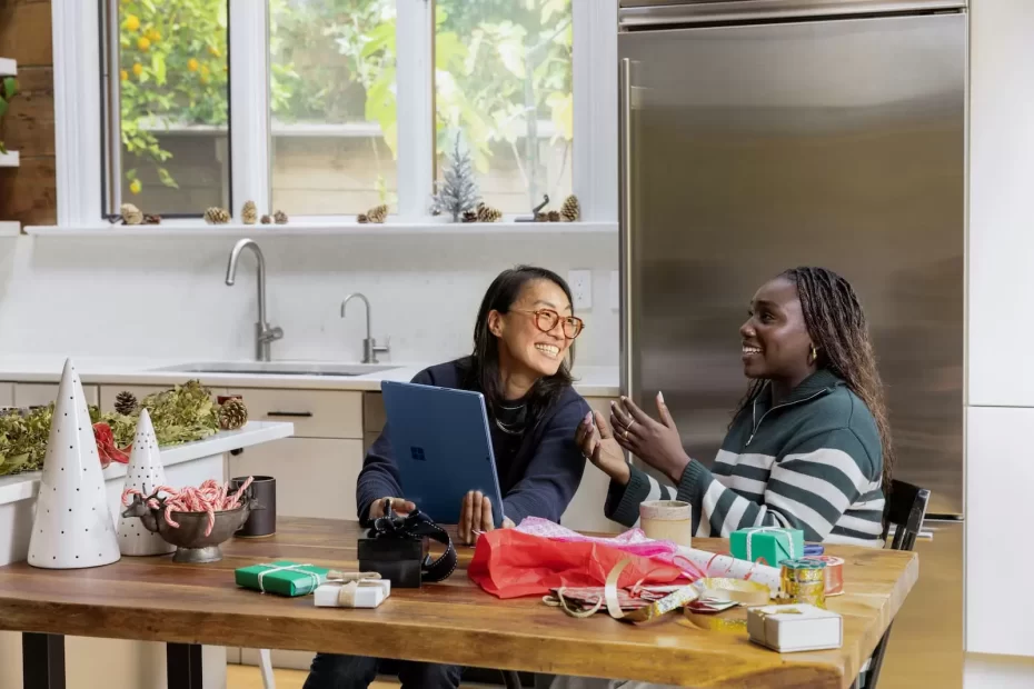 2 women sitting in the kitchen table. The woman to the left is showing the woman to the right something funny on a tablet